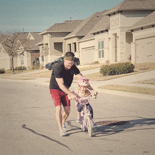 TJ running alongside his daughter as she rides a small pink bike down a neighborhood street